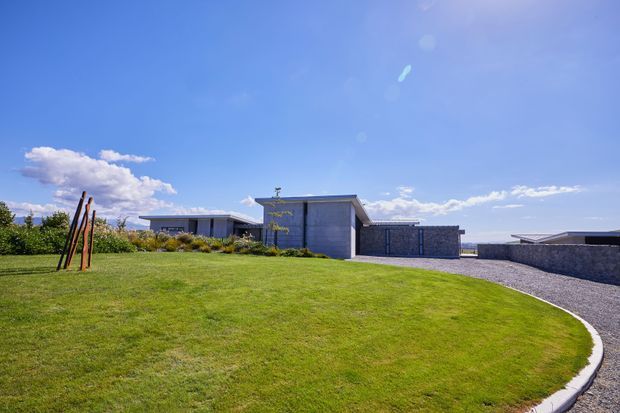 Wide exterior view of a low-slung modern residence set behind a broad manicured lawn. The building features flat rooflines, strong horizontal composition, dark grey masonry or concrete walls, extensive glazing, and a curved paved or gravel driveway/path along the foreground. A simple sculptural timber element stands in the lawn, with open sky and distant landscape beyond.