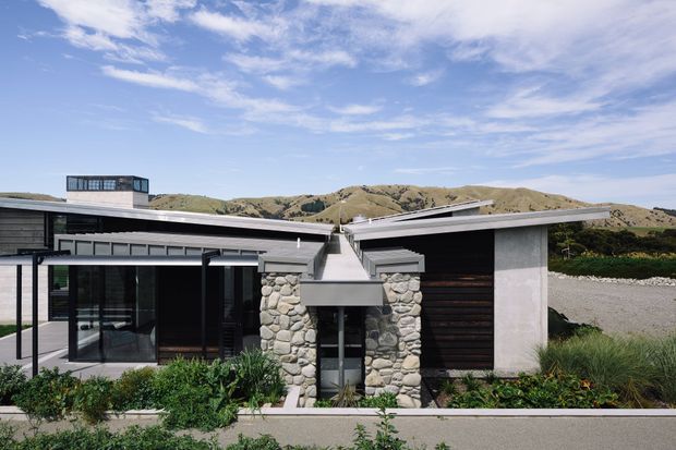 Exterior view of a low-slung contemporary house set against rolling hills. The building features broad flat or very low-pitched roof planes, extensive dark-framed glazing, dark horizontal timber cladding, pale rendered walls, and a prominent natural rounded-stone entry surround. Native-style landscaping and a gravel driveway sit in the foreground and beside the house.