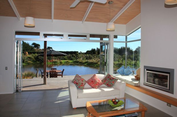 Bright contemporary living room opening onto a deck with expansive floor-to-ceiling glazing and views over a landscaped pond. The room features a white sofa with colourful patterned cushions, a glass-topped timber coffee table, tiled flooring, a built-in fireplace, and a warm timber-lined ceiling with exposed white beams and ceiling fans.