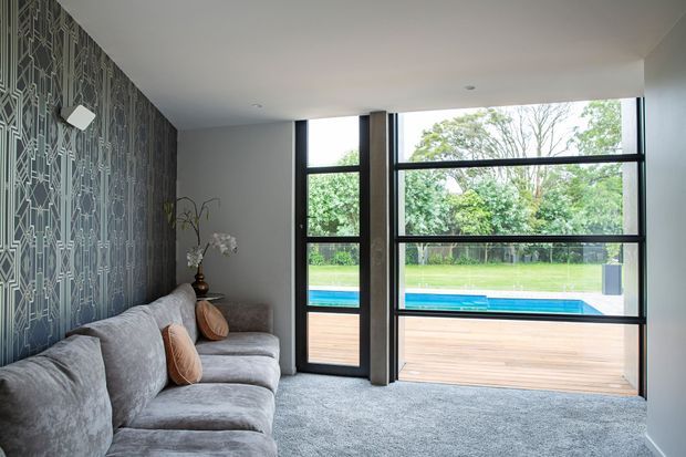 A contemporary living room with a large grey sectional sofa, textured grey carpet, and a dark geometric feature wall. Floor-to-ceiling black-framed glazing opens the room visually to a timber deck and swimming pool, with abundant greenery beyond. The room has a low, clean-lined ceiling and a restrained, refined palette.
