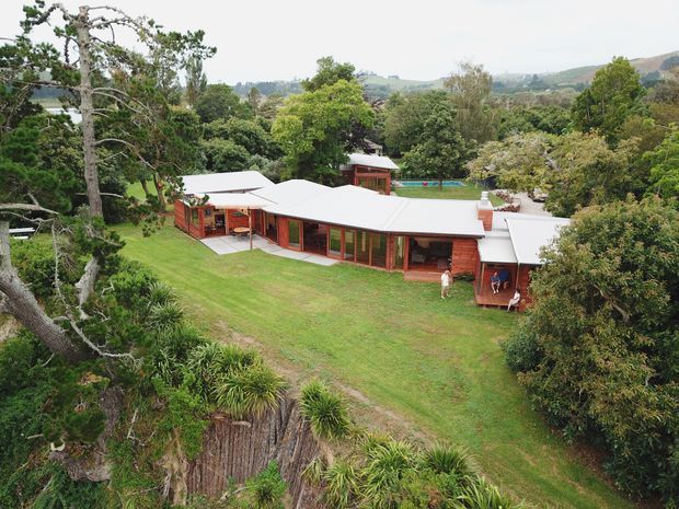 Aerial view of a low, sprawling single-storey residential building set within a lush landscaped, sloping site. The home has multiple connected wings with pale metal roofing, extensive reddish-brown timber-framed glazing, and dark timber wall sections. A broad lawn surrounds the building, with dense mature vegetation, a swimming pool visible behind the house, and rolling hills in the distance.
