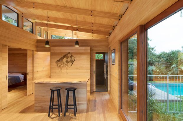 Warm timber-lined open-plan interior with a compact built-in kitchen or serving counter and two stools in the foreground. A bedroom is visible through a doorway on the left, while a narrow hallway leads toward a bathroom or utility area at the rear. Extensive timber wall, ceiling and floor finishes, exposed joinery, pendant lights, and full-height glazing overlook a swimming pool and landscaped garden.