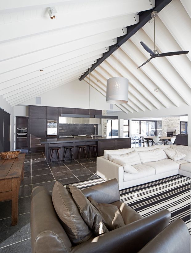Bright open-plan interior combining a dark timber kitchen with island seating and a spacious living area. The room has a dramatic white exposed-rafter pitched ceiling, dark structural beam, suspended pendant lights, ceiling fan, large windows, dark tiled flooring, and contrasting white and brown leather sofas.