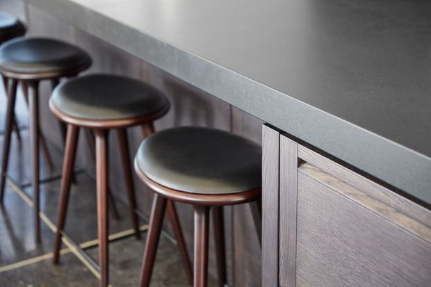 Close-up of a contemporary kitchen or bar counter with a dark grey benchtop, timber cabinetry, vertical wall panelling, and a row of dark upholstered stools with timber legs.
