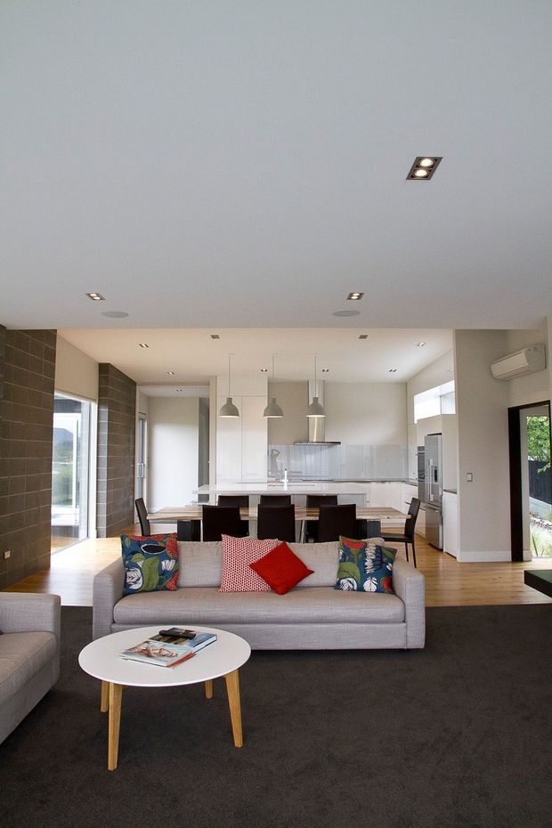 Open-plan contemporary living and dining area with a modern white kitchen in the background. The room features a grey sectional sofa, colourful patterned cushions, a red accent cushion, round white coffee table, dark carpet, timber flooring, pendant lights, recessed ceiling lighting, tiled feature walls, and large glazed openings.
