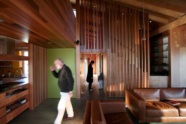 Warm, open-plan residential interior featuring extensive timber wall and ceiling lining, built-in timber cabinetry, a compact kitchen at left, and a living area with brown leather seating. A tall vertical timber-slat screen forms a dramatic partition around the hallway, with shelving and a green door visible beyond. The space has a high ceiling and strong natural timber character.