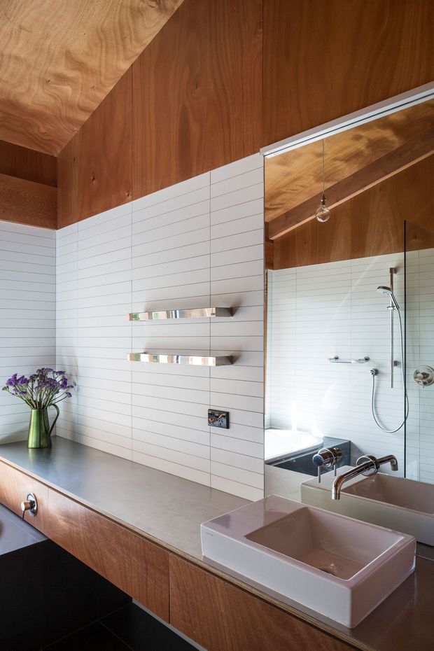 A refined contemporary bathroom with a long timber vanity, integrated countertop, pale pink vessel basin, large wall mirror, and wall-mounted chrome fixtures. The walls are clad in white horizontal rectangular tiles, while warm timber continues across the ceiling and built-in cabinetry. A shower area and freestanding-style bath are visible in the mirror reflection. The room has a warm, sophisticated material palette with clean linear detailing.