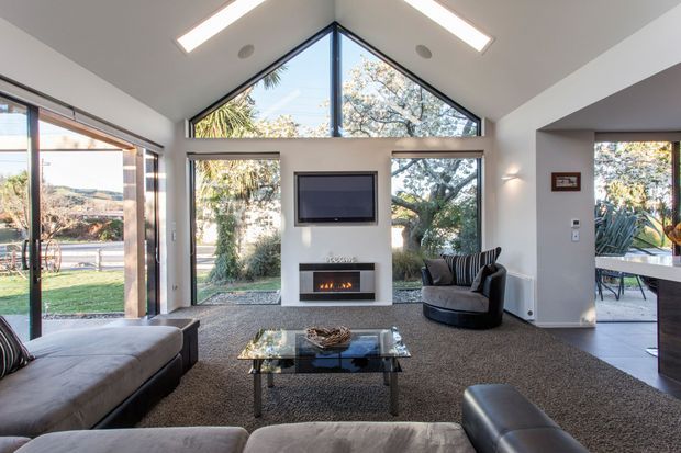 Bright contemporary living room with a vaulted ceiling and dramatic triangular clerestory glazing. Full-height windows and sliding doors open the room to a garden and surrounding trees, while a central fireplace with wall-mounted television forms the focal point. The room includes large upholstered sofas, a dark lounge chair, a glass coffee table, and recessed linear ceiling lights.