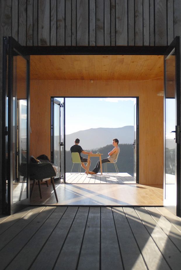 A compact timber-lined interior opens through large black-framed glazed folding or pivot doors onto a small deck with two people seated at a table, overlooking a mountainous landscape. The space features warm plywood wall and ceiling finishes, dark timber floorboards, extensive glazing, and strong sunlight creating long shadows. A dark upholstered lounge chair sits to the left. The architecture has a simple, cabin-like indoor-outdoor connection and an elevated scenic setting.