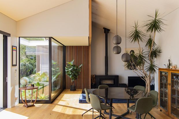 A bright, open-plan mid-century-inspired living and dining area with warm timber flooring, full-height glazing, vertical timber screening, sculptural pendant lights, a black fireplace, and indoor plants. The room opens visually to a planted outdoor courtyard and is furnished with a round dining table and upholstered chairs.