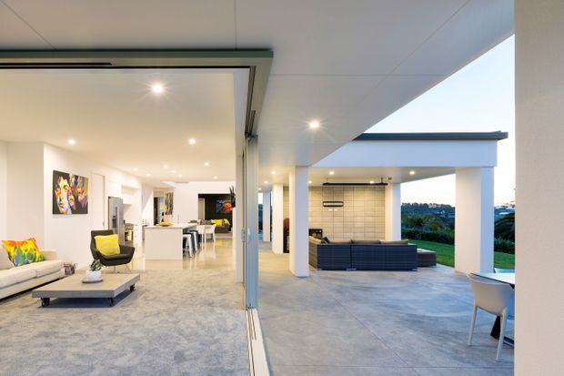 A bright contemporary open-plan living area opening through large sliding glass doors to a covered outdoor entertaining space. The interior includes a pale sectional sofa, low coffee table, colourful artwork and a dining area, while the exterior features a concrete-paved terrace, white structural columns, an outdoor sofa, built-in barbecue or kitchen zone, and landscaped views beyond.