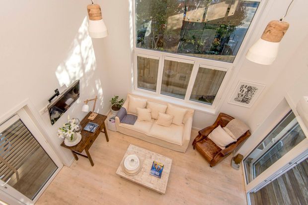 A bright double-height living room viewed from above, featuring pale timber flooring, a cream sofa with cushions, a brown leather armchair, a marble or light stone coffee table, a small timber side table, pendant lights, and extensive windows and glazing. The room has a calm, contemporary interior with a strong sense of vertical space and natural light.