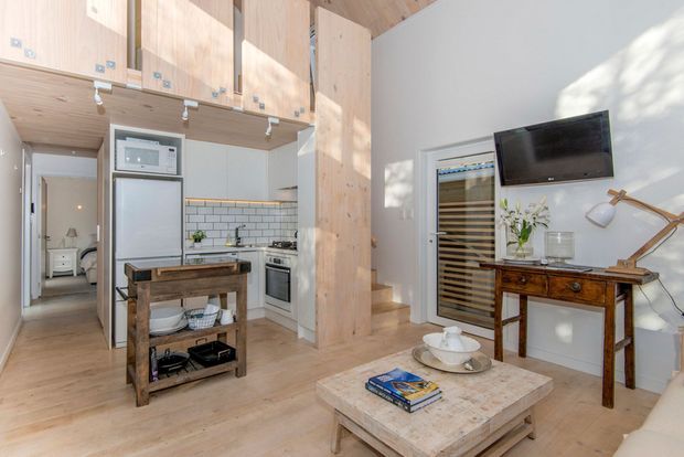 Bright compact open-plan living and kitchen interior with a loft-like layout. Pale timber cabinetry and ceiling panels frame a white tiled kitchen with stainless-steel appliances, while a rustic wooden island cart, vintage console table, wall-mounted television, wine refrigerator, and pale stone coffee table furnish the room. Sunlight creates strong patterned shadows on the white wall, giving the space an airy, warm character.