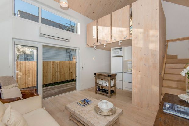 Compact open-plan living room with a light timber-lined ceiling, pale timber flooring, and a small white kitchen tucked beneath a loft-style mezzanine. Tall timber cabinetry and a timber stair enclosure define the right side of the room, while large glazed doors and high windows bring in natural light. A neutral sofa, armchair, coffee table, and compact rustic island trolley furnish the space. The architecture has a warm, efficient small-home character with strong vertical storage and loft circulation.