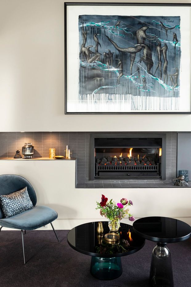 A refined living room vignette featuring a contemporary fireplace clad in dark grey horizontal tiles, a pale built-in media or display cabinet, framed abstract artwork, a sculptural blue lounge chair, and glossy black round coffee tables. The room has charcoal carpet, neutral walls, reflective surfaces, and colourful floral accents, creating a sophisticated, moody atmosphere.