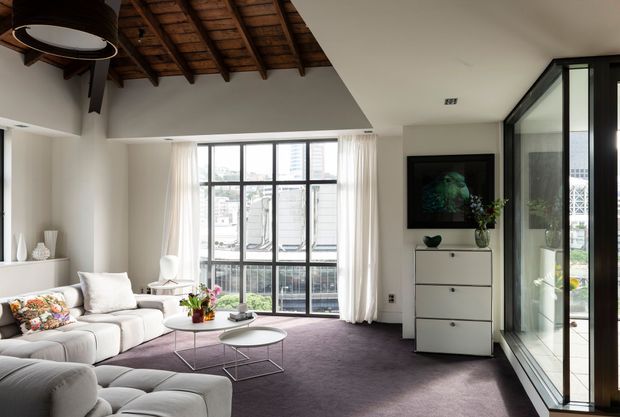 Bright contemporary living room with a large sectional sofa, nested round coffee tables, pale built-in shelving, a tall chest of drawers, and floor-to-ceiling black-framed glazing. The room features a deep grey-purple carpet, white walls and curtains, and a distinctive exposed timber-rafter ceiling section with dark structural beams.