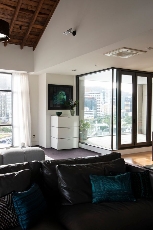 A bright contemporary living room with a dramatic exposed timber vaulted ceiling detail, white walls, dark-framed floor-to-ceiling glazing, and panoramic urban views. The room includes a large black leather sectional sofa, a white chest of drawers, framed artwork, and a small balcony or terrace visible beyond the glazing.