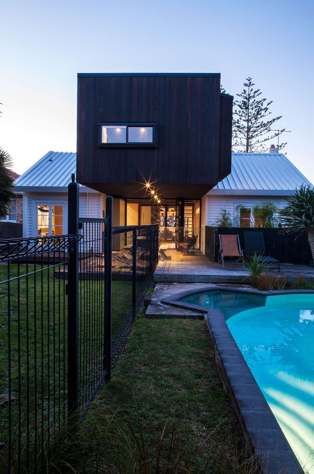 Twilight exterior view of a renovated or extended suburban house with a prominent dark timber-clad upper-level box projecting over a ground-floor entry and deck. The lower level has pale weatherboard walls and light metal pitched roofs, with black metal fencing, a timber deck, landscaped planting, and a swimming pool in the foreground. Warm interior and exterior lighting creates a welcoming contrast against the cool blue evening sky.