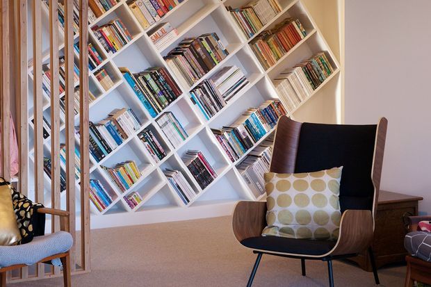 A warm, eclectic reading nook featuring a large white built-in bookshelf arranged in a striking diagonal grid and filled with books. In front of it is a sculptural mid-century-style lounge chair with wooden arms, a patterned cushion, and dark metal legs. A timber-framed hanging chair and side furniture add to the inviting, layered interior.