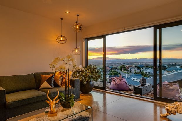 Warm, contemporary living room with a dark upholstered sectional sofa, glass coffee table, indoor plants and decorative pendant lighting. Full-height sliding glass doors open onto a balcony or terrace with outdoor seating, overlooking a suburban coastal or waterfront landscape at sunset. Polished light flooring and expansive glazing create a relaxed, elevated outlook.