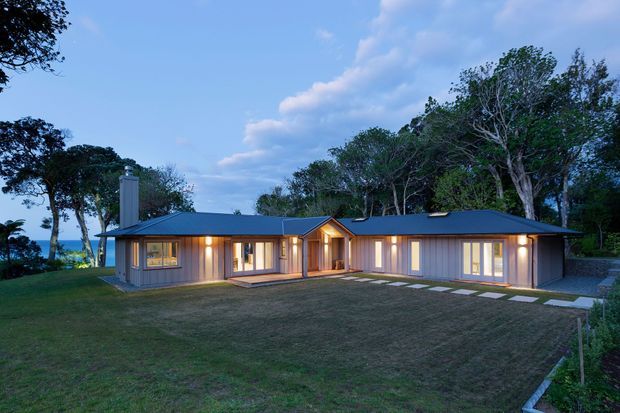 Single-storey contemporary timber-clad residence with a low-pitched dark roof, broad glazed openings, warm interior lighting, and a connected winged floor plan. The house sits within a landscaped, wooded setting with a large lawn and stepping-stone path.