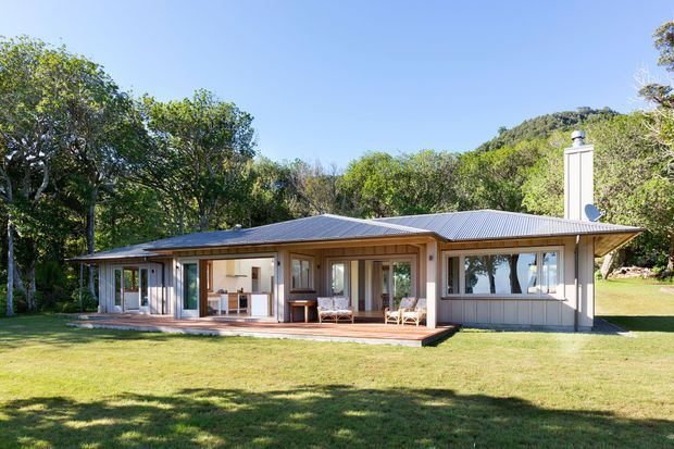 Single-storey pavilion-style house set in a leafy landscaped setting, with a low-pitched corrugated metal roof, light vertical weatherboard cladding, extensive glazed doors and windows, and a broad timber deck opening onto a lawn. The architecture has a relaxed contemporary character with strong indoor-outdoor connection and a prominent chimney.