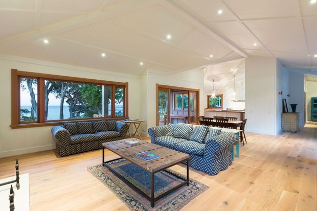 Bright open-plan living room with timber flooring, patterned upholstered sofas, a rustic rectangular coffee table, and a vintage rug. Large timber-framed windows provide views toward water and trees, while the adjoining dining area and kitchen are visible beyond. The room has white paneled walls and ceiling with recessed lighting, creating a relaxed coastal character.