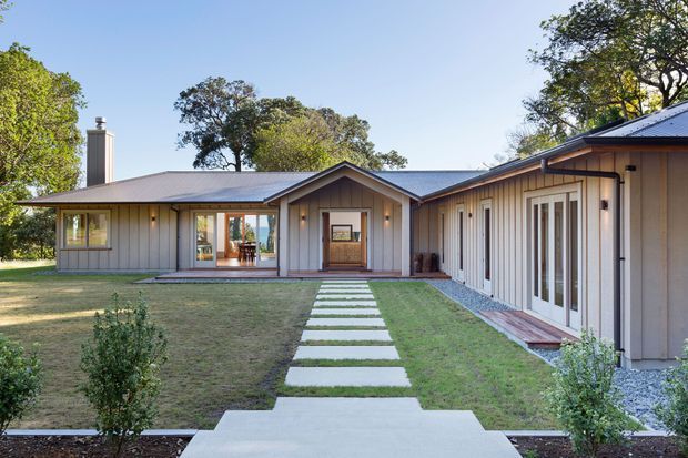 Single-storey low-profile residential building arranged around a broad central lawn and straight stepping-stone walkway. The facade features grey vertical board cladding, dark pitched metal roofs with gabled entry volumes, white-framed glazed doors and windows, and a covered veranda-like transition between wings. Mature trees and landscaped planting surround the house.