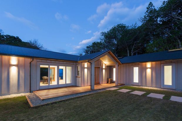 Single-storey contemporary residential building viewed from a landscaped courtyard at dusk. The L-shaped structure has dark pitched metal roofs, vertically clad light timber or composite walls, warm exterior wall lighting, large glazed doors opening to a timber deck, and a sheltered timber-lined entry porch. Stepping-stone paving crosses the lawn toward the entrance, with trees beyond.