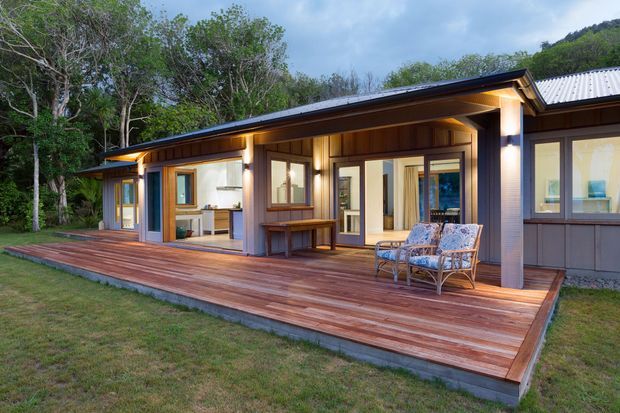 Single-storey contemporary house set among dense greenery, viewed from a broad timber deck. The facade combines dark grey horizontal cladding, timber-framed glazing, exposed timber soffits, and a corrugated metal roof. Large sliding doors open toward the deck, revealing a bright interior, while outdoor lounge chairs and a built-in bench occupy the covered deck.