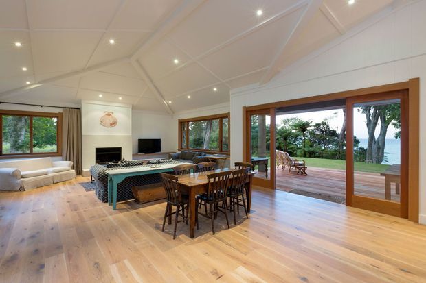 Spacious open-plan living and dining room with pale timber flooring, white walls, and a high vaulted ceiling with exposed painted beams. The room includes a dining table, lounge seating, fireplace, television, and patterned upholstered furniture. Large timber-framed glazed doors open onto a broad timber deck overlooking mature trees and a water view.