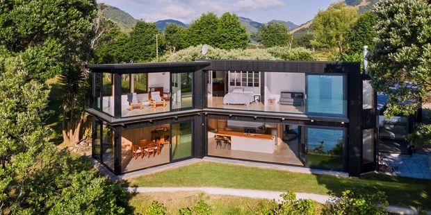 A two-storey contemporary glass-walled residence set within a lush landscaped site, with a dark structural frame, extensive floor-to-ceiling glazing, and mountain views. Living, dining, and kitchen areas are visible on the lower level, while bedrooms and a blue-toned bathroom are visible upstairs.