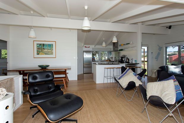 Open-plan living and kitchen interior with mid-century-inspired lounge furniture, a compact island kitchen, timber flooring, white wall panelling, exposed ceiling beams, and pendant lighting. The room is bright and relaxed, with large windows connecting to the outdoor setting.
