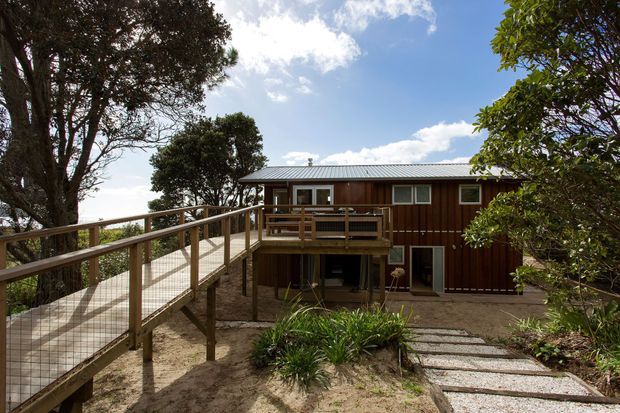 Exterior view of a raised two-storey dwelling set among mature trees, with dark timber vertical cladding, a low-pitched metal roof, elevated timber deck, and an accessible ramp leading to the upper level. Stepping stones and landscaped planting surround the lower entry area.