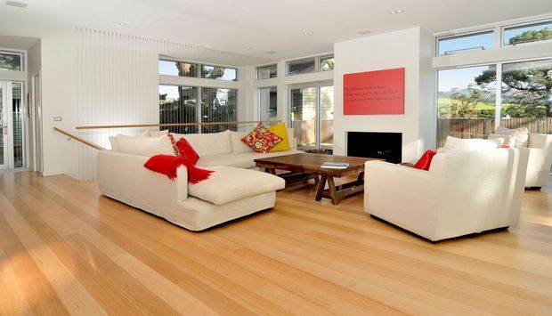 Bright, spacious contemporary living room with pale timber flooring, white upholstered sofas and armchairs, extensive glazing, a minimalist fireplace wall, and colourful red and patterned accents. The room opens toward a stair or circulation area and is filled with natural light.