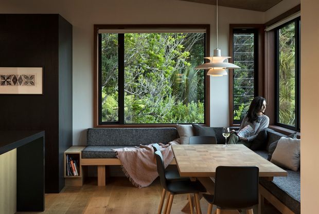 A warm, compact open-plan living and dining area with a built-in corner banquette, charcoal upholstered seating, a timber dining table, and black dining chairs. Large timber-framed windows overlook dense greenery, while a sculptural pendant light hangs above the table. Dark built-in cabinetry and a fireplace or media wall occupy the left side, with timber flooring and a timber ceiling creating a cohesive natural palette.