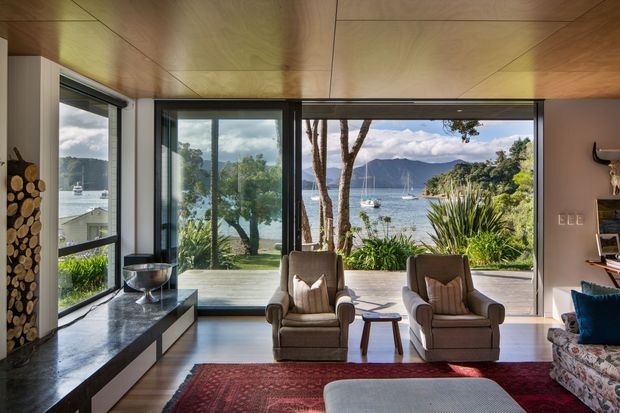 A refined living room overlooking a sheltered waterfront inlet, with expansive floor-to-ceiling glazing, timber-lined ceiling, dark-framed windows, a stone or dark tiled fireplace hearth, two upholstered lounge chairs, a patterned red rug and a partial sofa. The view includes water, boats, trees and mountainous terrain beyond.