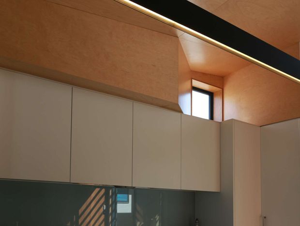 A close architectural interior view showing full-height pale grey built-in cabinetry beneath a warm timber-lined ceiling and upper wall surfaces. A narrow high-set window brings in daylight, while a dark reflective splashback or wall surface is visible below the cabinetry. A long black linear ceiling element with integrated lighting creates a strong horizontal accent. The image is minimalist, warm, and highly focused on fixed joinery and architectural finishes.