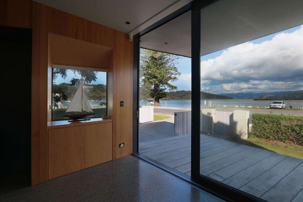 Interior view of a contemporary waterfront living space opening onto a paved deck and landscaped frontage. The room features warm timber wall panelling and built-in cabinetry, dark-framed floor-to-ceiling glazing, and a polished grey floor. A large sliding glass opening provides views toward a calm body of water and distant hills, with a model sailboat displayed in the joinery.