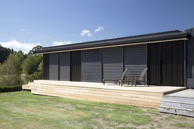 Single-storey modern dark-clad building viewed from the exterior, with a broad low-pitched roof, full-width louvred and glazed-looking facade elements, and a raised pale timber deck with steps. Two outdoor chairs and a small table sit on the deck; grass and trees surround the structure.