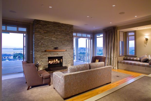 A spacious, refined living room centred on a substantial stacked-stone fireplace with an inset fire. Upholstered sofas and armchairs face the fireplace, while floor-to-ceiling windows and glazed doors open to a balcony with expansive water and landscape views. Built-in window seating, layered curtains, recessed lighting, and a warm timber floor border create an elegant, comfortable interior.