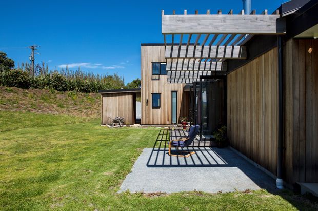 Exterior view of a contemporary timber-clad residence opening onto a concrete patio and broad lawn. A prominent timber pergola projects over the patio, casting strong linear shadows, while dark-framed glazing and vertical wood cladding define the facade. The setting appears suburban or semi-rural, with planted embankments and clear sky in the background.