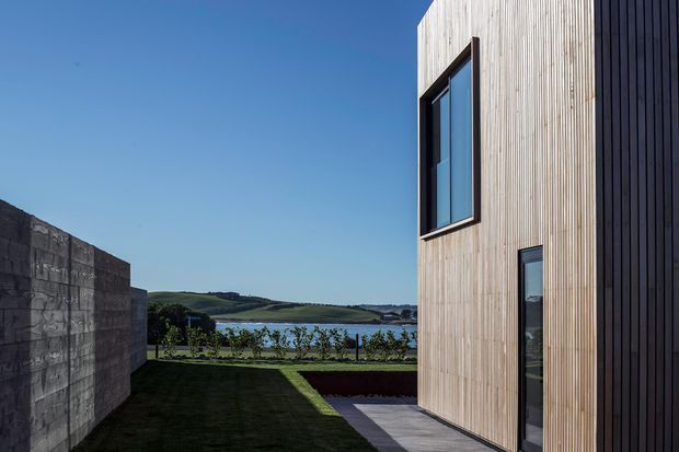 Exterior architectural photograph of a contemporary multi-level building clad in pale vertical timber battens, with a darker vertical-clad side wall and dark-framed windows. A paved path and landscaped lawn lead toward a body of water, with rolling green countryside visible beyond under a clear blue sky.