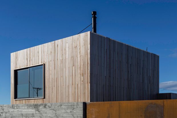 Exterior view of a compact, cubic modern building clad in closely spaced vertical timber battens. The simple flat-roofed volume sits behind a rough concrete retaining wall and a rusted corten-like metal fence, with a large dark-framed window and a black chimney projecting above the roof. Clear blue sky and strong sunlight emphasize the crisp geometric form.
