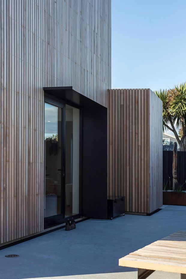 Exterior view of a contemporary building with tall vertical timber screening and cladding, dark recessed glazing and a projecting black entry canopy. A paved outdoor area and timber deck occupy the foreground, with established planting at the edge of the property.