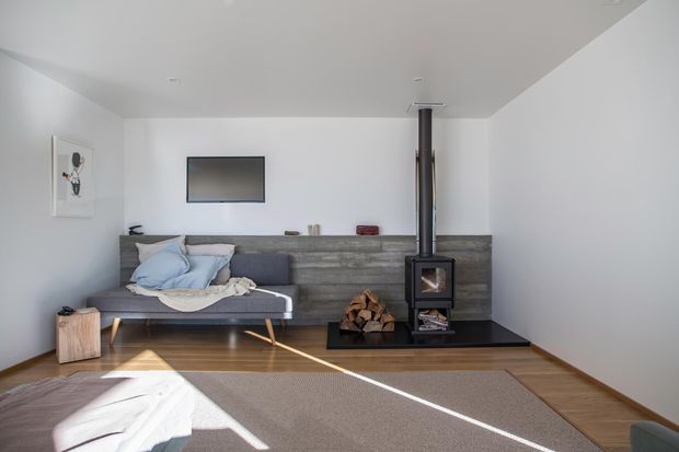 A bright, minimalist living room with a grey sofa, wall-mounted television, timber flooring, and a freestanding black wood-burning stove set against a horizontal grey feature wall with stacked firewood beneath. White walls and ceiling, sparse furnishings, and strong sunlight create a clean contemporary atmosphere.