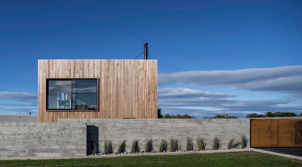 Exterior view of a compact contemporary building with a flat roof, vertical timber-clad upper volume, large dark-framed window, and a long raw concrete boundary wall with a rust-toned metal gate. Minimal landscaping and a deep blue sky emphasize the clean, rectilinear composition.