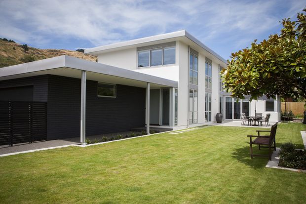 Exterior view of a contemporary two-storey home with a crisp white flat roof, extensive floor-to-ceiling glazing, dark horizontal cladding, and a covered carport or entry canopy. The house opens onto a neatly landscaped lawn with a paved terrace and outdoor dining furniture.
