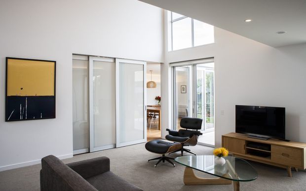 Bright contemporary living room with a high ceiling and clerestory glazing. The room features white walls, grey carpet, a dark grey sofa, a black leather lounge chair with ottoman, a glass coffee table, and a timber media console. Sliding glass doors open toward a dining area and glazed outdoor connection. A large abstract black-and-mustard artwork adds a small accent.