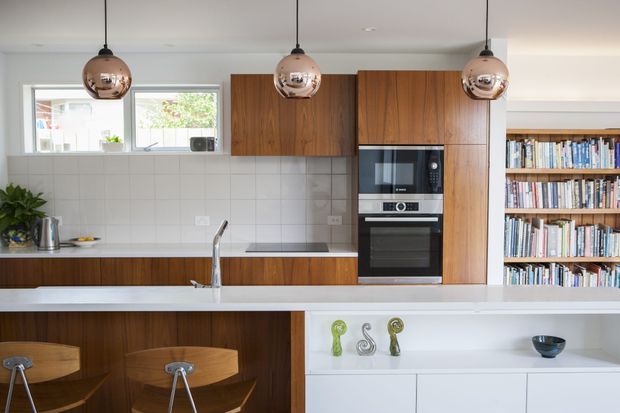 Bright contemporary kitchen with a long white island and integrated sink, timber cabinetry, white tiled splashback, built-in stainless-steel oven and microwave, and three copper-toned pendant lights. Timber bar stools sit at the island, while open white shelving and a large built-in bookcase occupy the right side of the room.